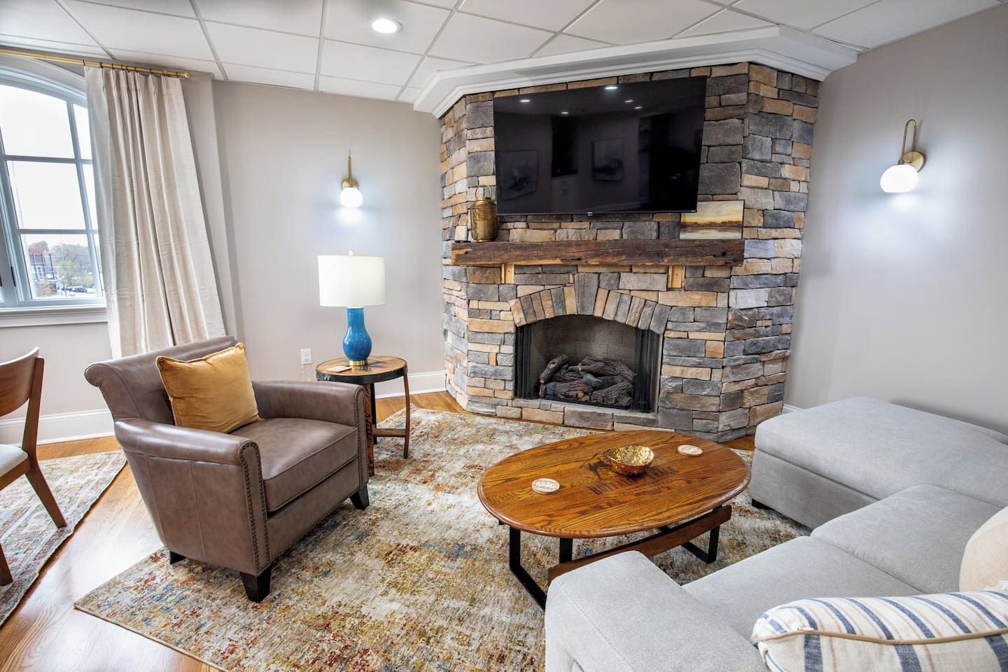 A living room with a stone fireplace, leather armchairs, a round coffee table, and a view through a large window.