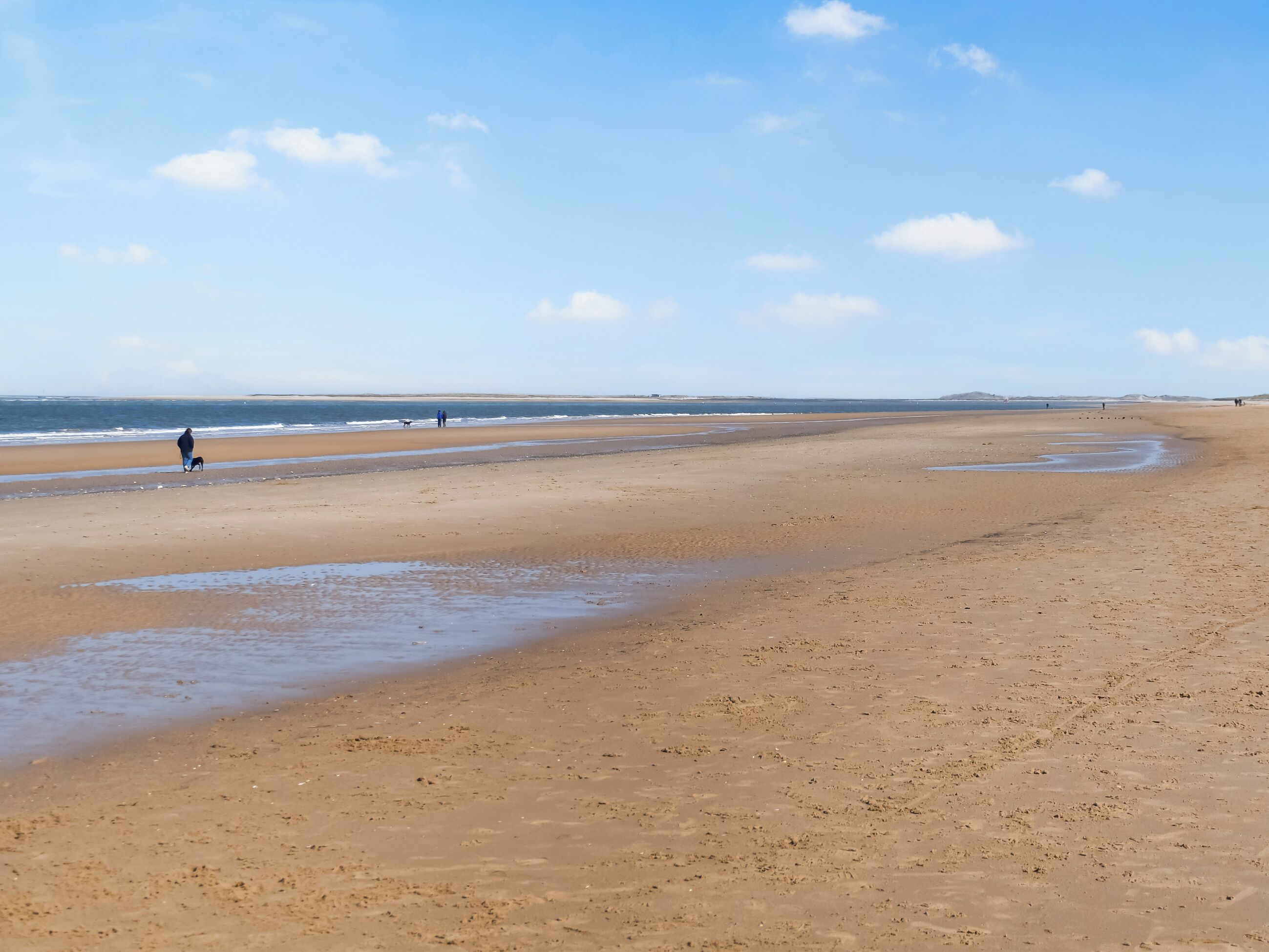 Un'ampia spiaggia sabbiosa con onde tranquille, cielo sereno e alcune persone che passeggiano.