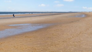 A wide sandy beach with gentle waves, a clear sky, and a few people walking.