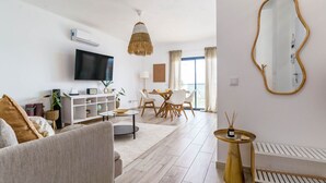 Een moderne woonkamer met een flatscreen televisie, een eethoek met een tafel en stoelen, en een groot raam met gordijnen.