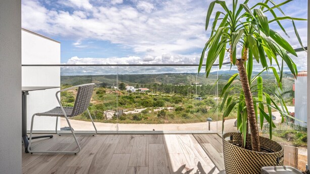 Een balkon met uitzicht op een glooiend landschap, een rieten stoel en een potplant.