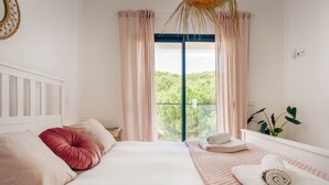 Een slaapkamer met een groot bed, twee kussens, een nachtkastje, een raam met gordijnen en uitzicht op groen.