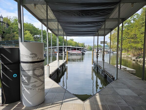 A covered dock area with a boat, a trash can, and a view of the water and surrounding greenery. - Margaritaville-Firepit- Private Dock-Tiki Bar-Free Golf (Osage Beach)
