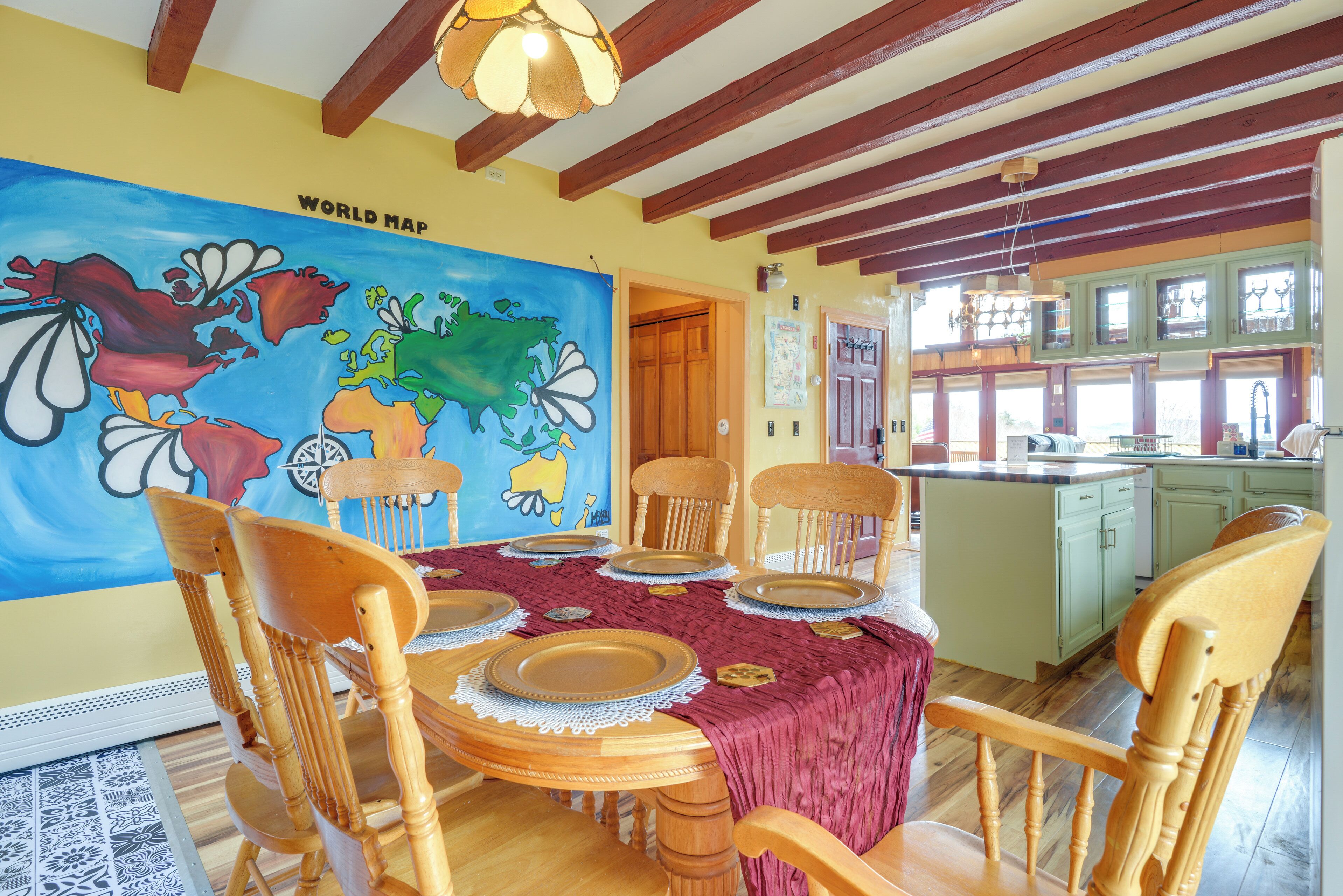 Un comedor con una mesa de madera dispuesta para seis personas, un mural de un mapa mundial en la pared y una cocina al fondo.
