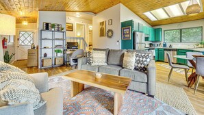 Een ruime zithoek met een houten plafond, een grijze bank met decoratieve kussens, een houten salontafel en een kleurrijk vloerkleed.