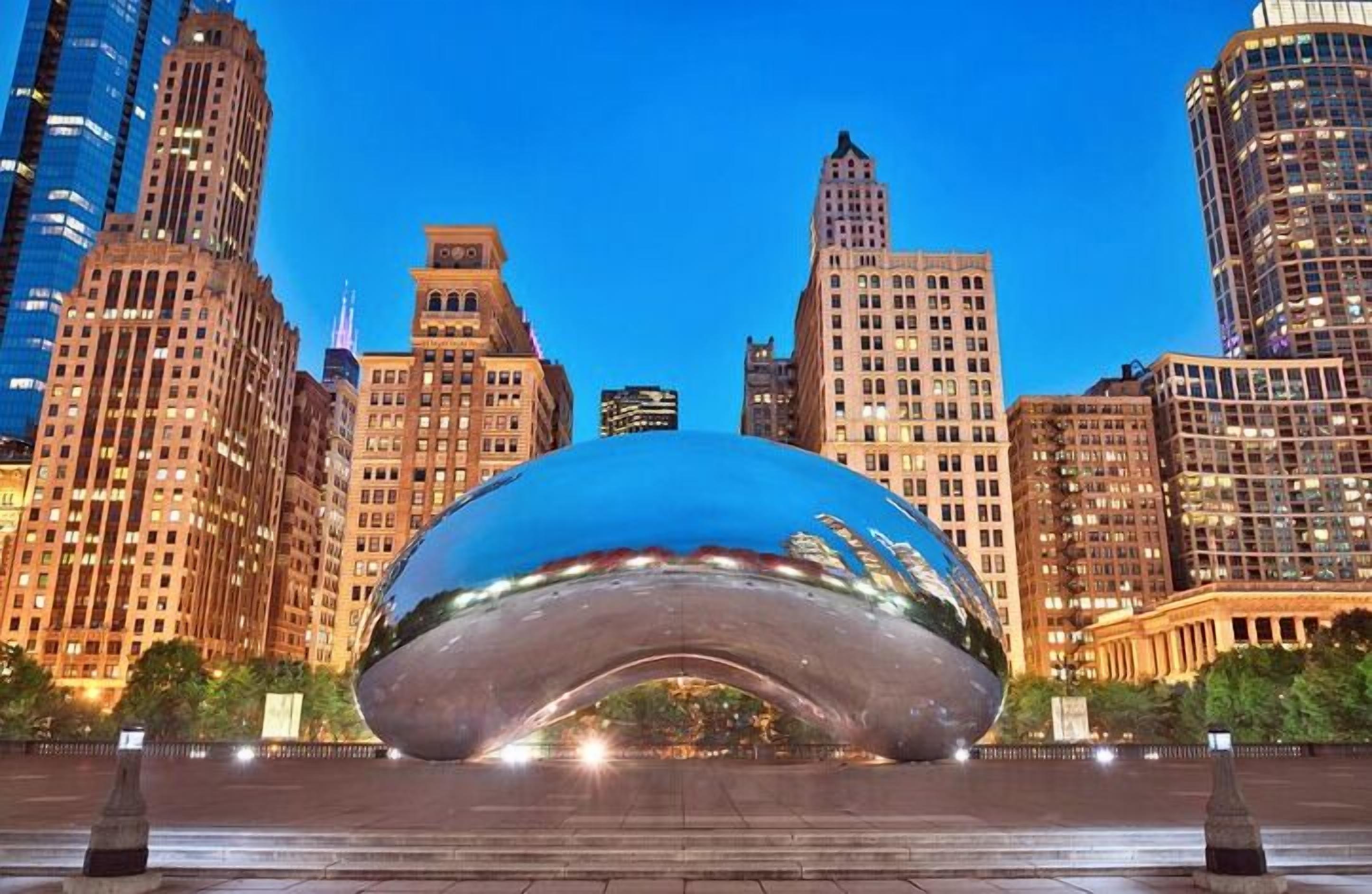 A large, reflective metallic sculpture in an urban setting with tall buildings in the background.