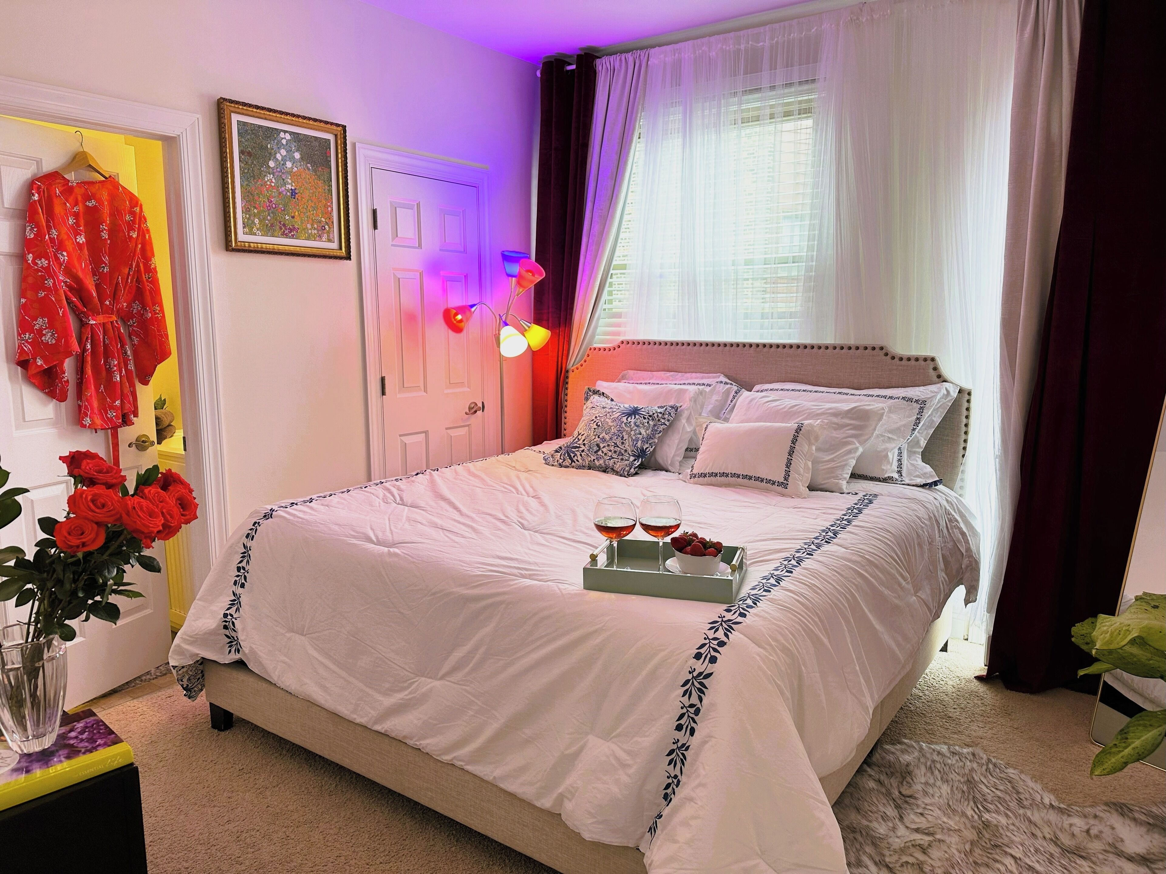 A neatly made bed with white bedding and blue floral trim, a bedside table with a tray holding two glasses of wine and a bowl of fruit, and a vase of red roses on the floor beside the bed.