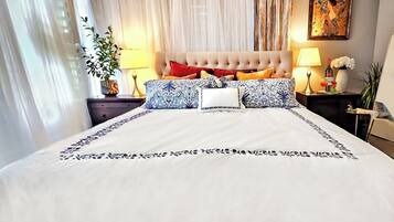 A neatly made bed with a white bedspread and decorative pillows, a tufted headboard, and a bedside table with a lamp and a vase of flowers.