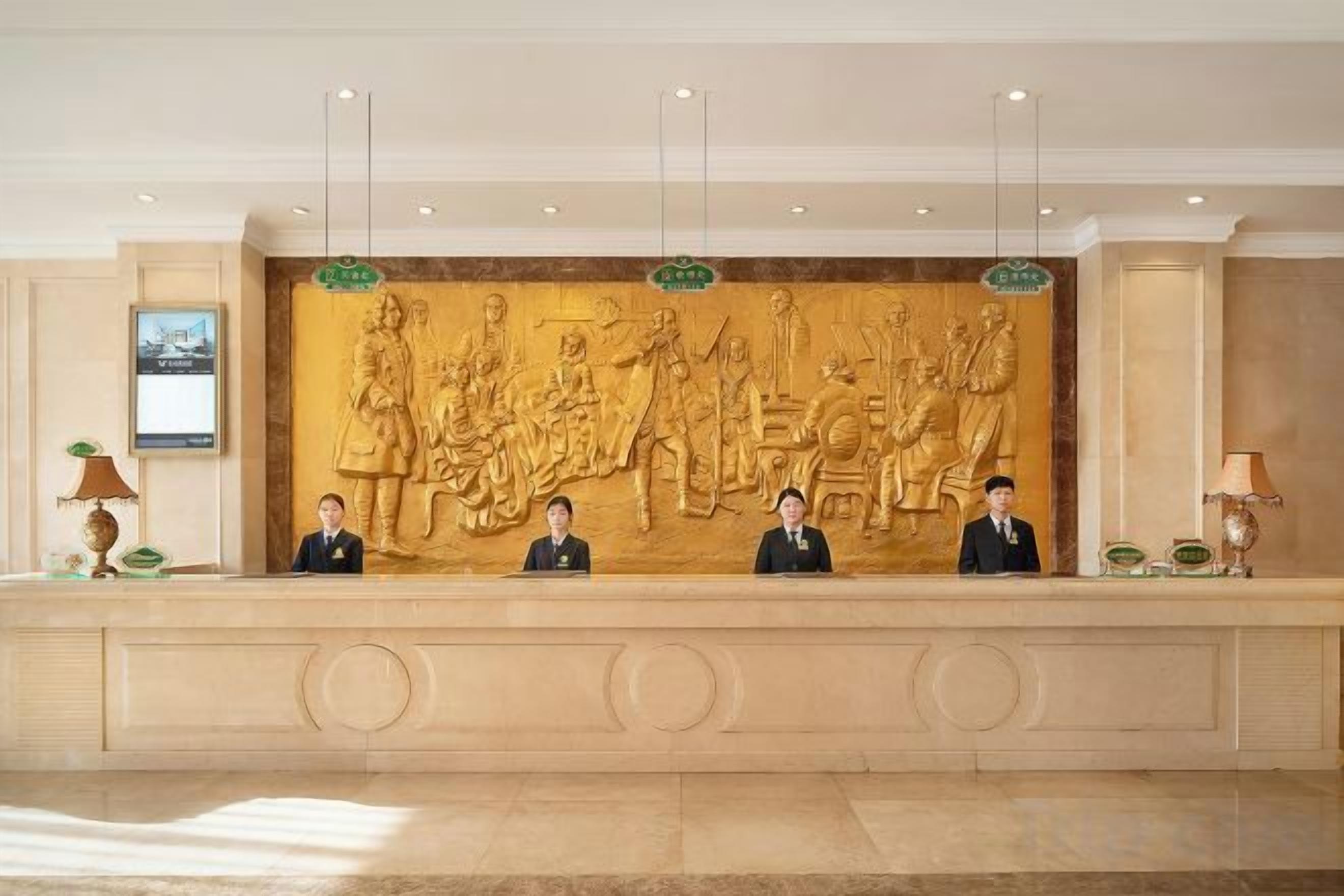 A reception desk with a large relief sculpture above it, three uniformed staff members seated behind the desk, and a framed picture on the wall to the left.