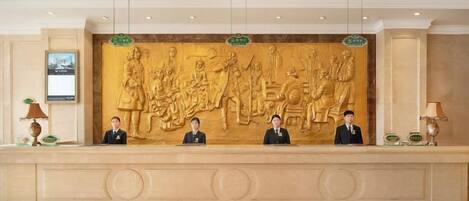 A reception desk with a large relief sculpture above it, three uniformed staff members seated behind the desk, and a framed picture on the wall to the left.