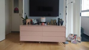 A living room with a flat-screen TV mounted on the wall, a pink dresser with various decorative items, and a framed photo display on the floor. - Lovely studio close to Champs Elysées (Paris)