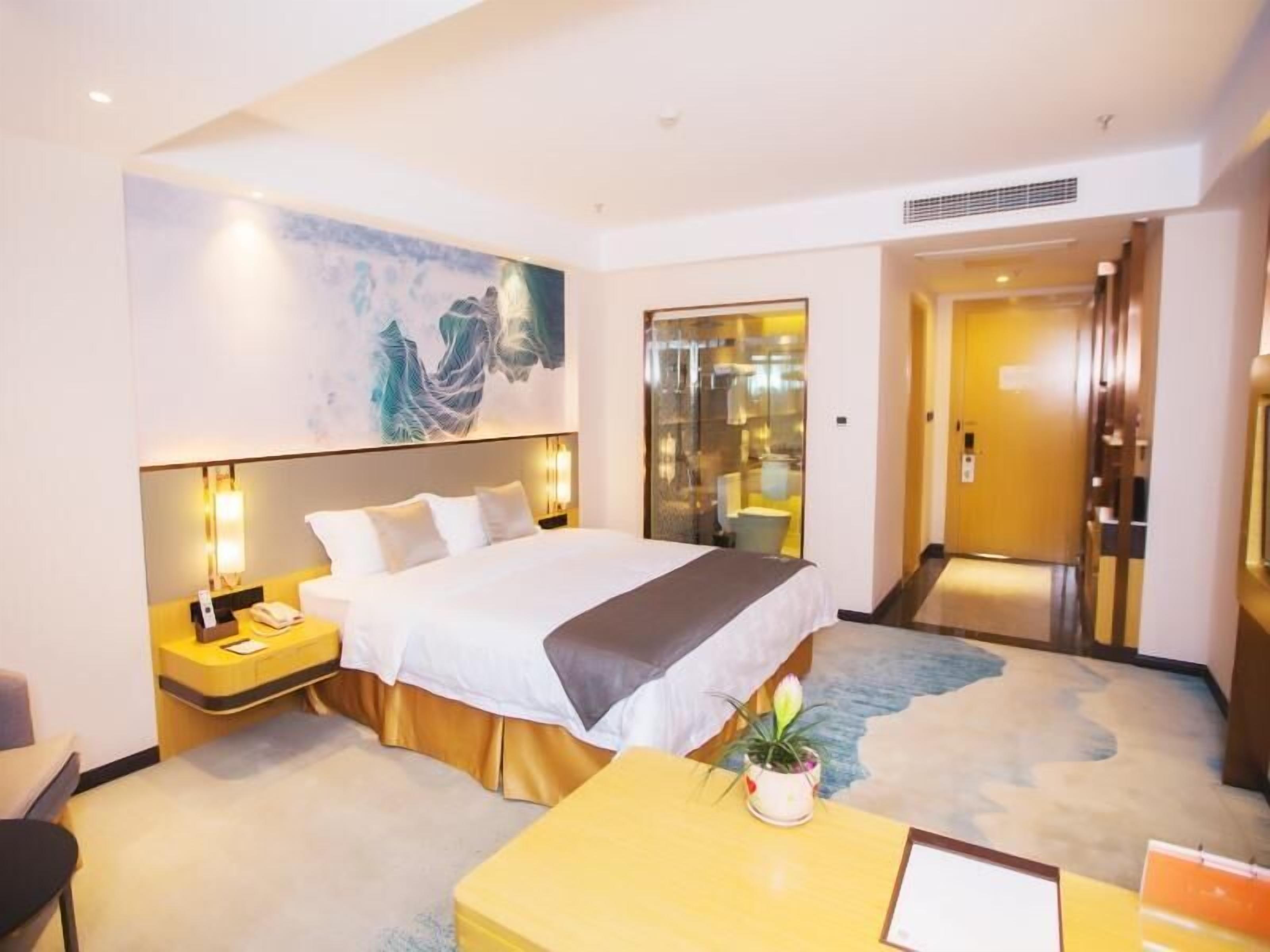 A hotel room with a large bed, bedside tables, a desk, a potted plant, and a painting on the wall.