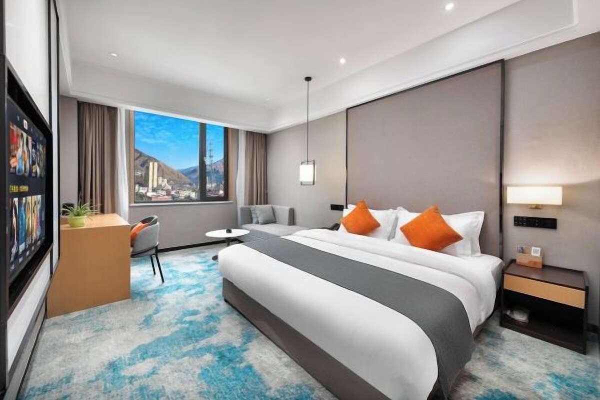 A modern hotel room with a large bed, a desk, a chair, a TV, and a view of a mountain landscape.