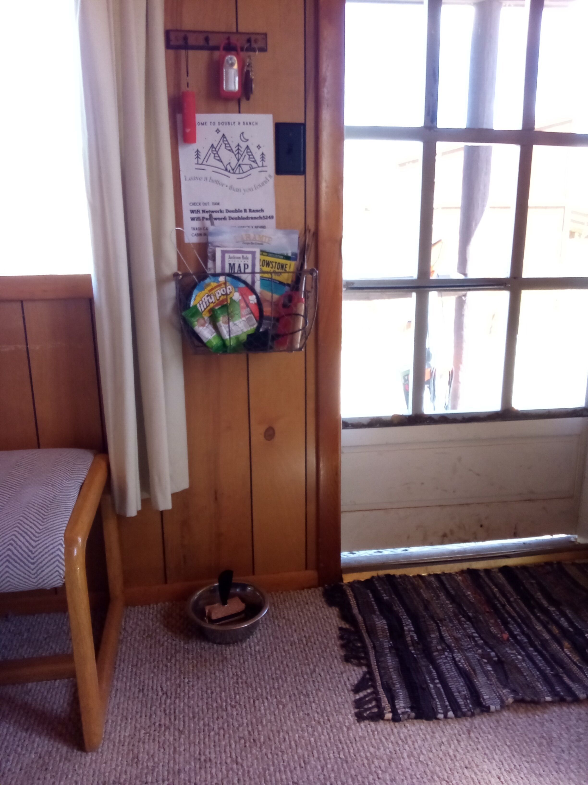 Un rincón de una habitación con una silla de madera, un tablón de anuncios con varios avisos, una ventana con barrotes y una alfombra en el suelo.
