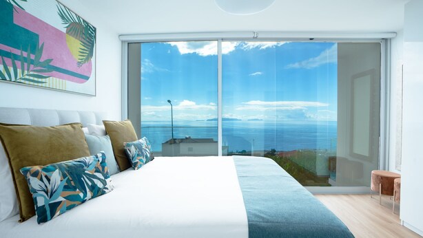 A bedroom with a large bed, a view of the sea through a sliding glass door, and a tropical-themed painting on the wall.