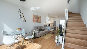A modern living room with a glass dining table, white chairs, a grey sofa, and a wooden staircase.