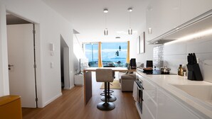 A modern kitchen with white cabinetry, a dining area with a high table and bar stools, and a view of the sea through large windows.