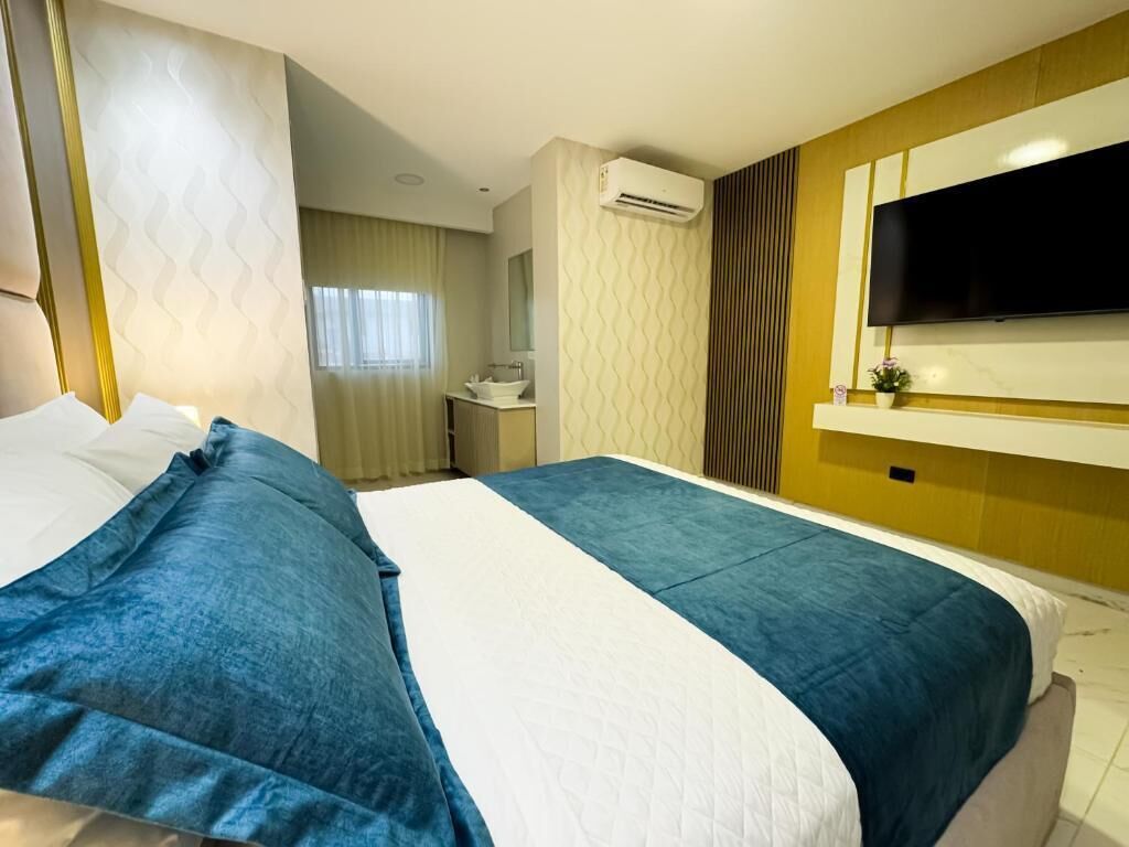 A hotel room with a large bed, a TV mounted on the wall, a bathroom with a sink and mirror, and an air conditioning unit.