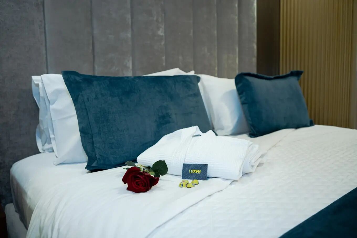 Una cama bien hecha con sábanas blancas, almohadas azules, una rosa roja y una tarjeta sobre la cama.