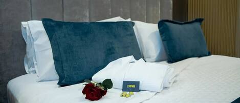 Una cama bien hecha con sábanas blancas, almohadas azules, una rosa roja y una tarjeta sobre la cama.