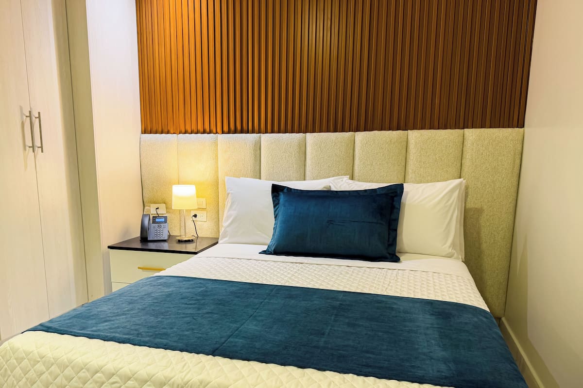 Quarto de hotel com uma cama grande, uma mesa de cabeceira com abajur e cabeceira de madeira.