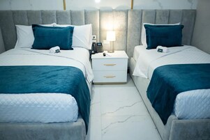 Habitación de hotel con dos camas, cada una con cubrecama azul y almohadas blancas. Hay una mesita de noche con una lámpara y un teléfono.