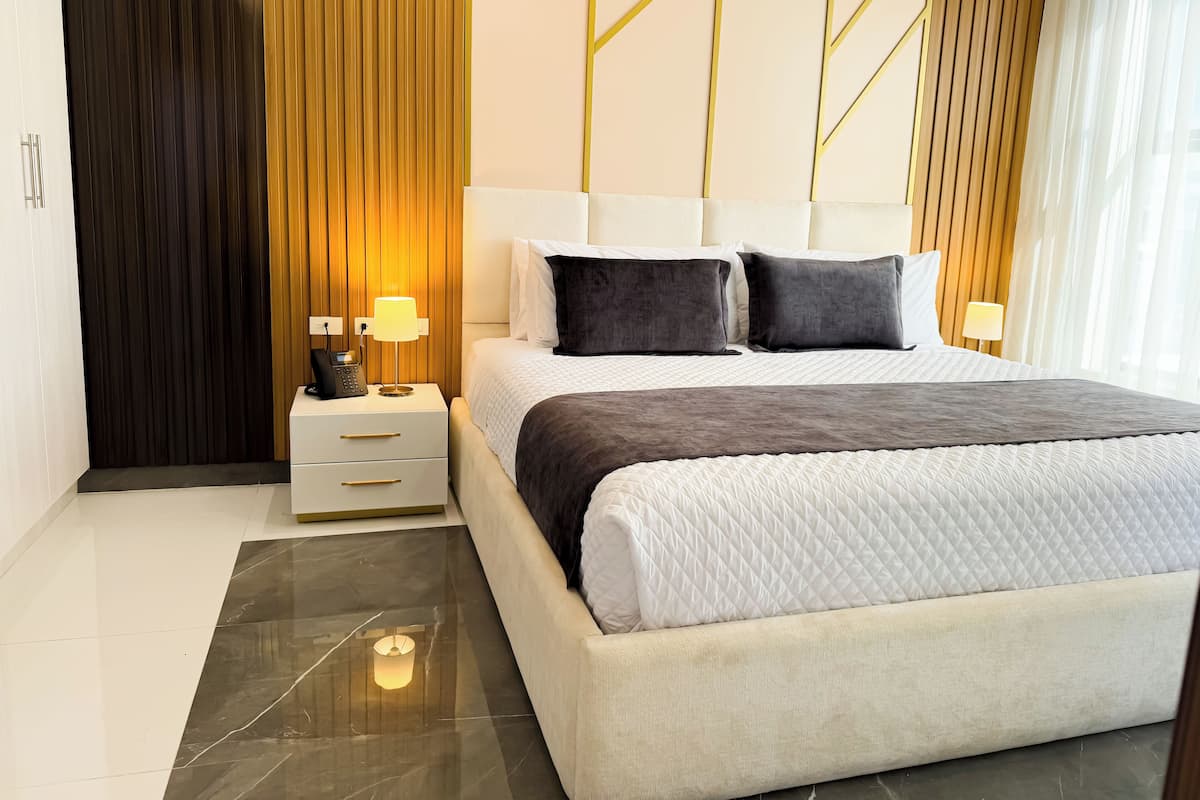 Quarto de hotel moderno com uma cama grande, mesa de cabeceira com abajur e parede de destaque em madeira.