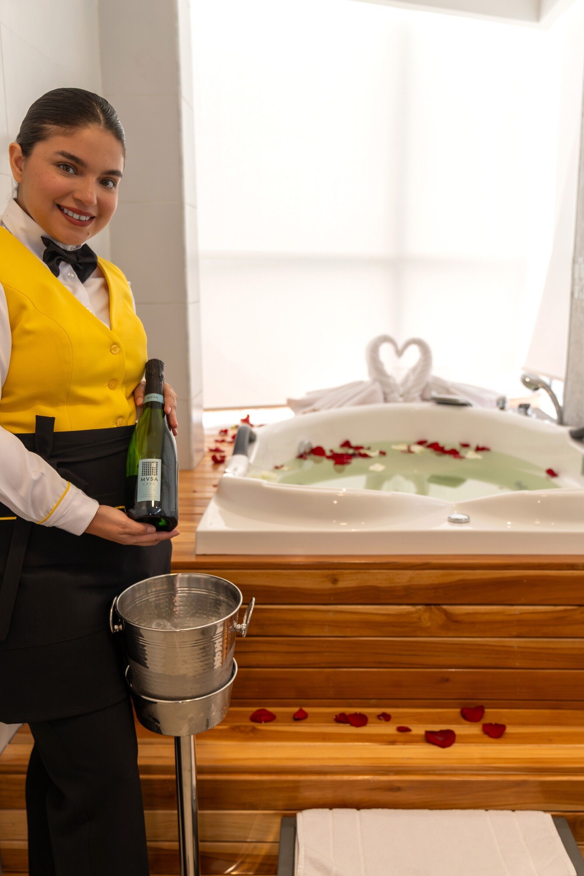 A person holding a bottle of wine next to a bathtub with rose petals.