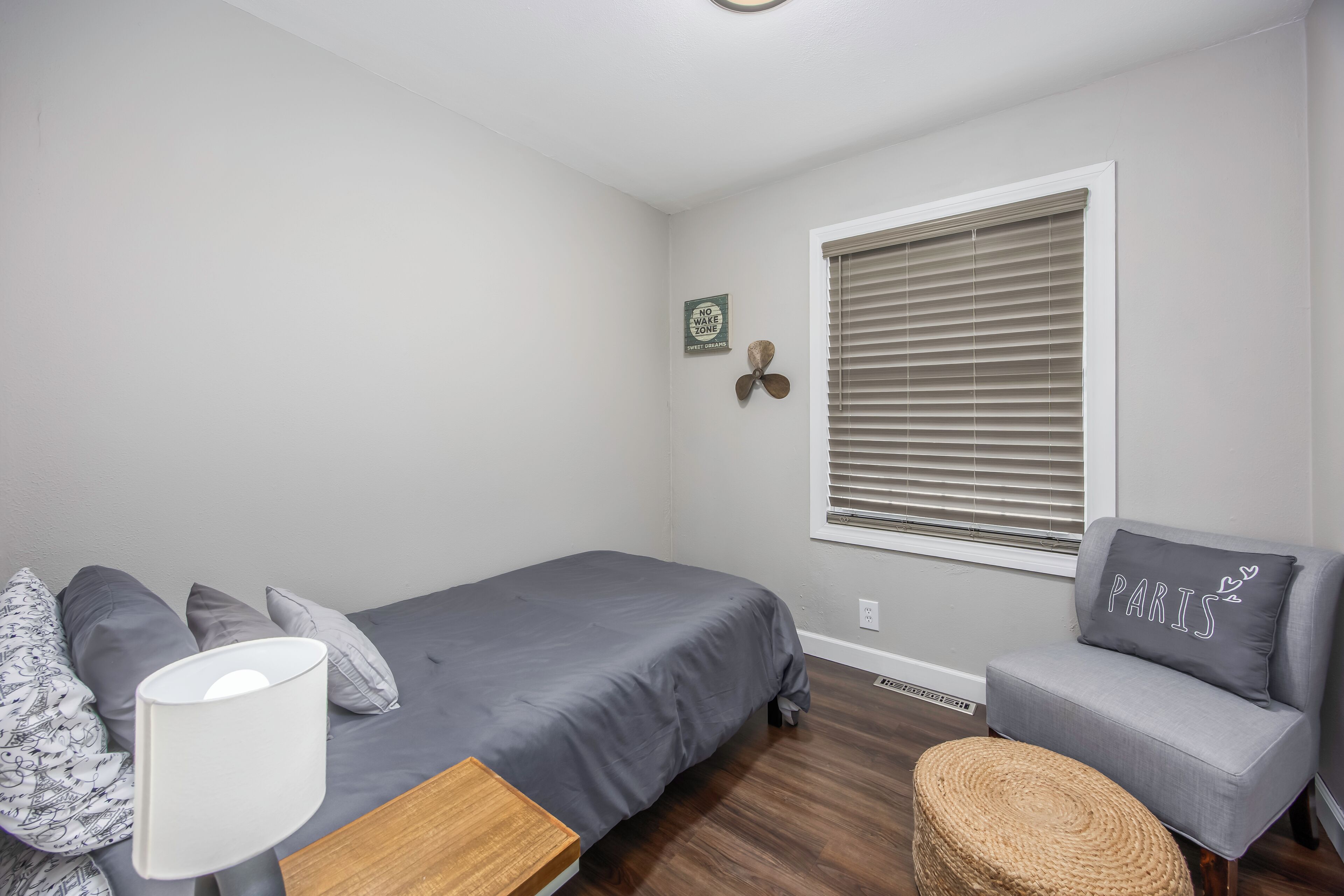 Une chambre à coucher comprenant un lit, une chaise, une lampe, une fenêtre avec des stores et un élément de décoration fixé au mur.