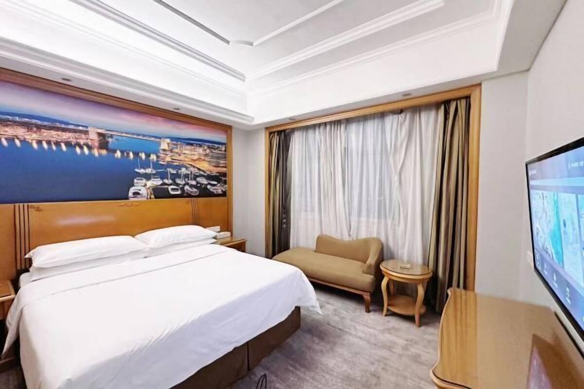A hotel room with a large bed, a sofa, a TV, and a painting of a harbor.