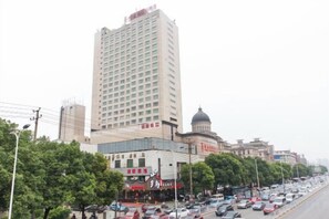 A multi-story hotel with a prominent building in the background, a busy street with cars, and trees lining the road. - Vienna International Hotel (Changsha Wanjiali Gaoqiao Market) (Changsha)