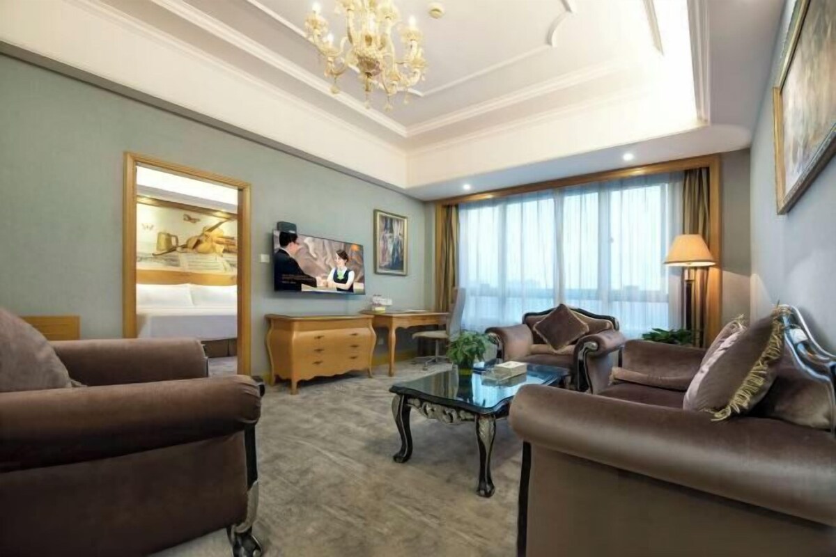 Quarto de hotel com uma cama grande, dois sofás de couro marrom, uma mesa de centro de vidro, uma escrivaninha, uma televisão e um candelabro.
