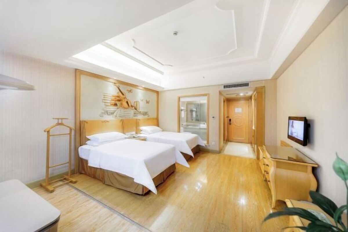 Quarto de hotel com duas camas, uma escrivaninha de madeira, televisão e um espelho com obras de arte.