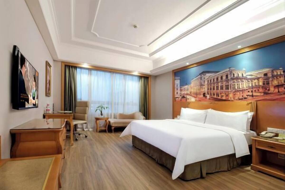 A hotel room with a large bed, a desk, a chair, a small table, and a large window with curtains.