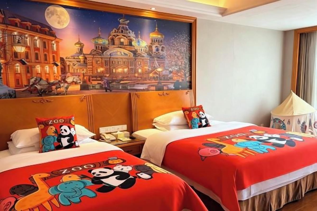A hotel room with two beds, each with a red blanket featuring cartoon pandas, a large painting of a cityscape, and a lamp on the right side.