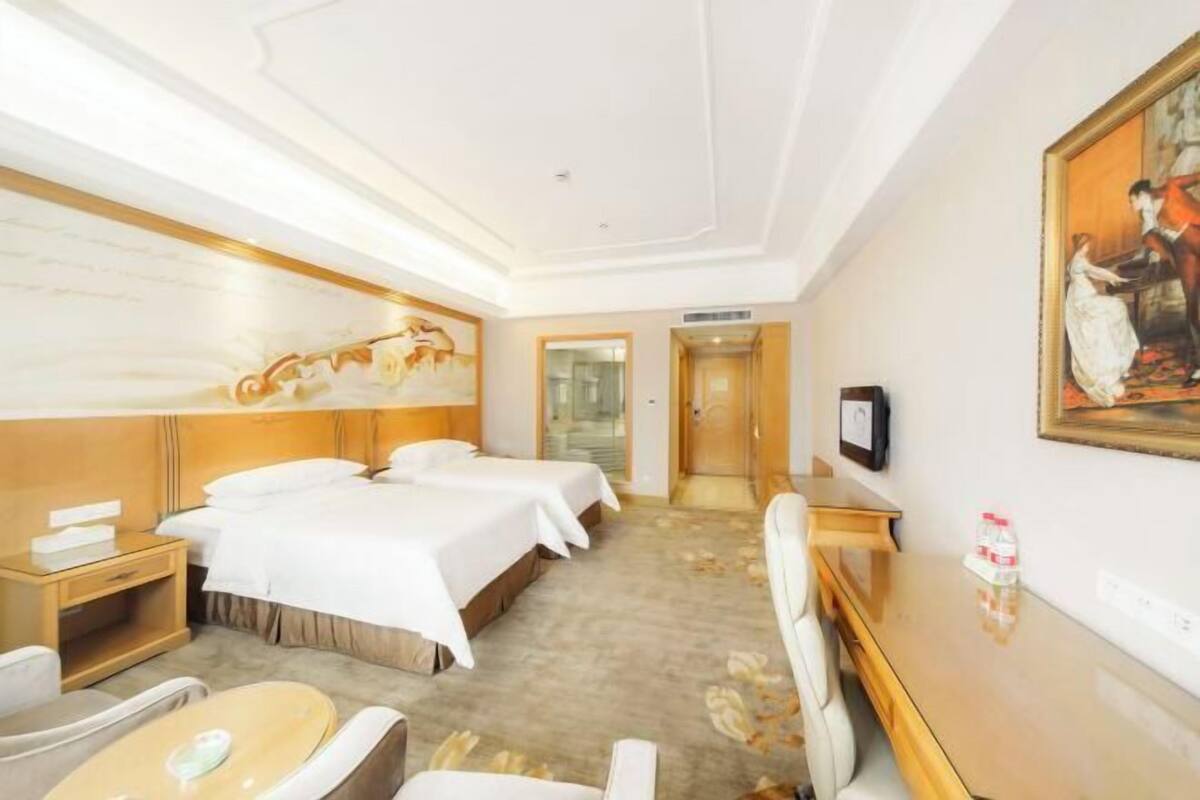 A hotel room with two beds, a desk, a chair, a TV, and a painting on the wall.