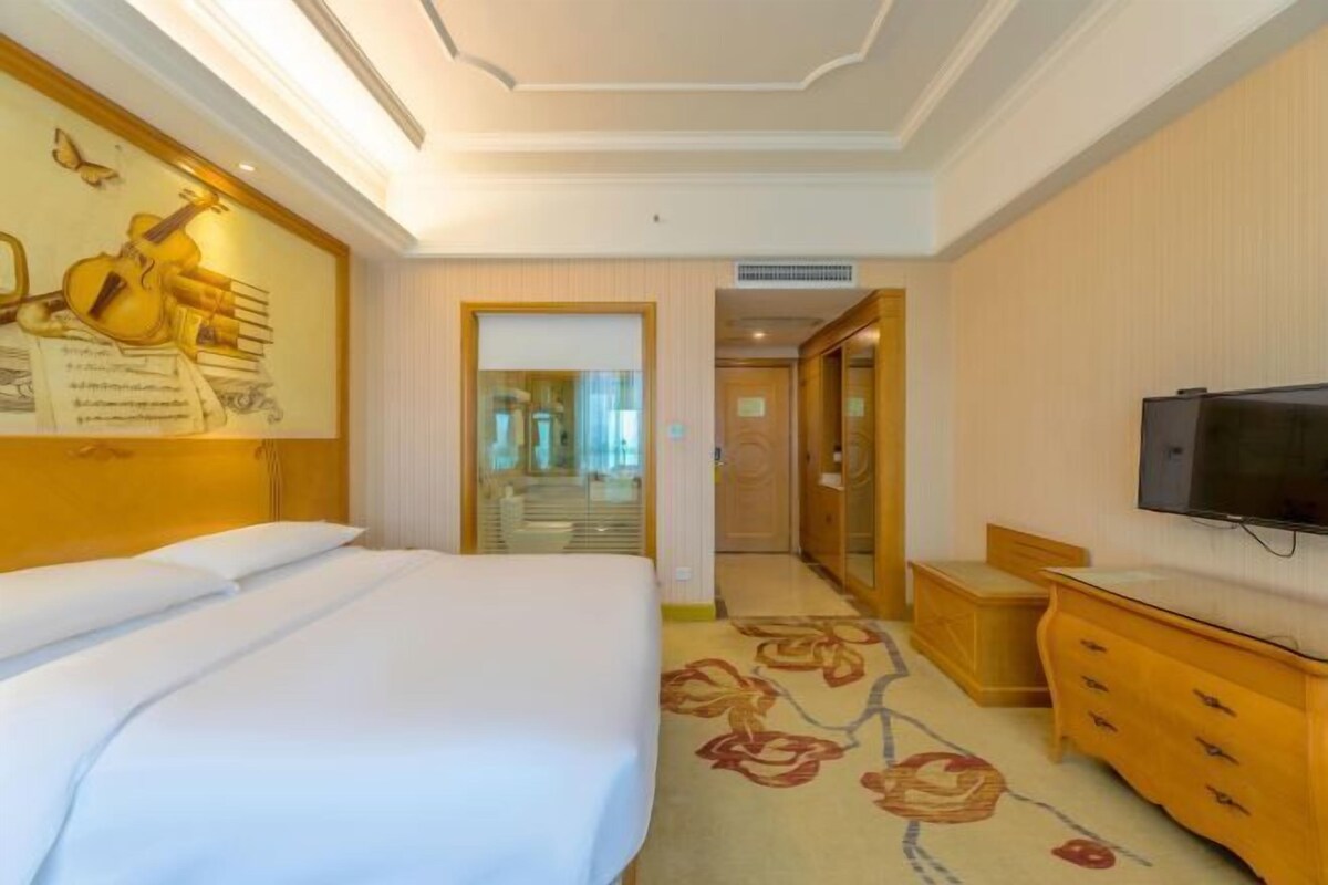 Quarto de hotel com uma cama, televisão, cómoda de madeira, tapete floral e uma parede com instrumento musical e partitura.