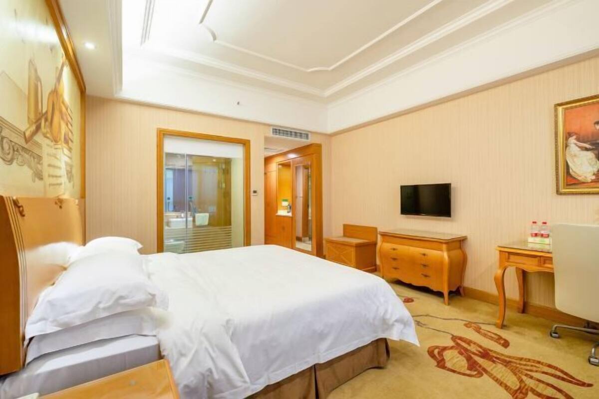 A hotel room with a bed, a desk, a chair, a television, and a painting on the wall.
