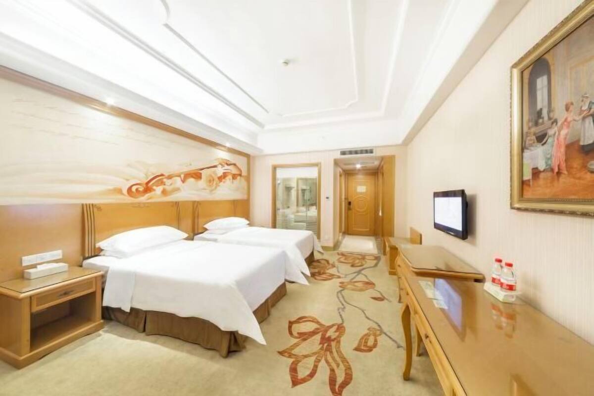 A hotel room with two beds, a desk, a television, and a painting on the wall.