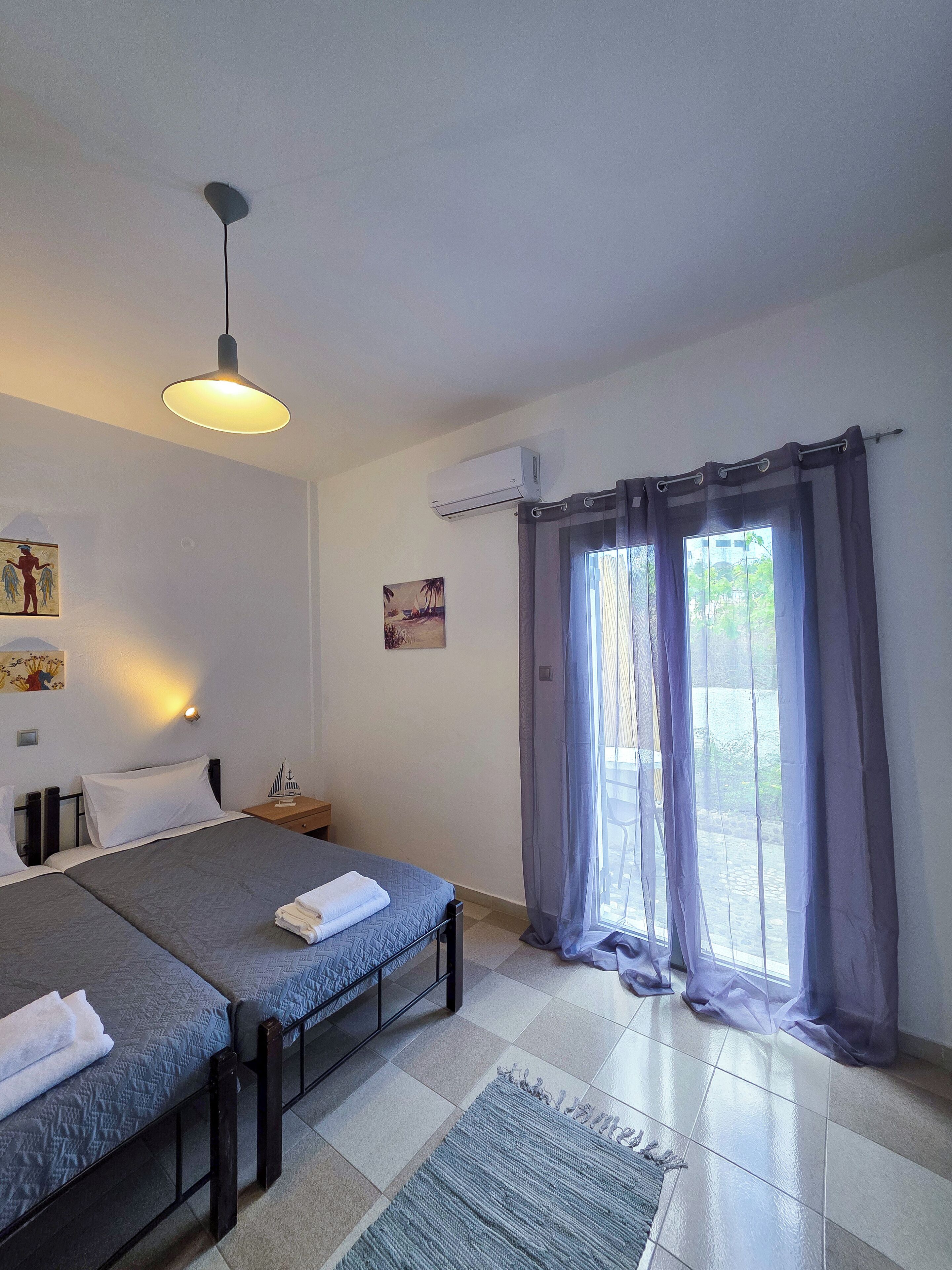 A room with two beds, a ceiling light, an air conditioner, a window with curtains, and a small table with a decorative item.