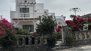A white two-story house with a balcony, surrounded by a stone wall and flowering plants. - Calypso Heaven (Santorini)