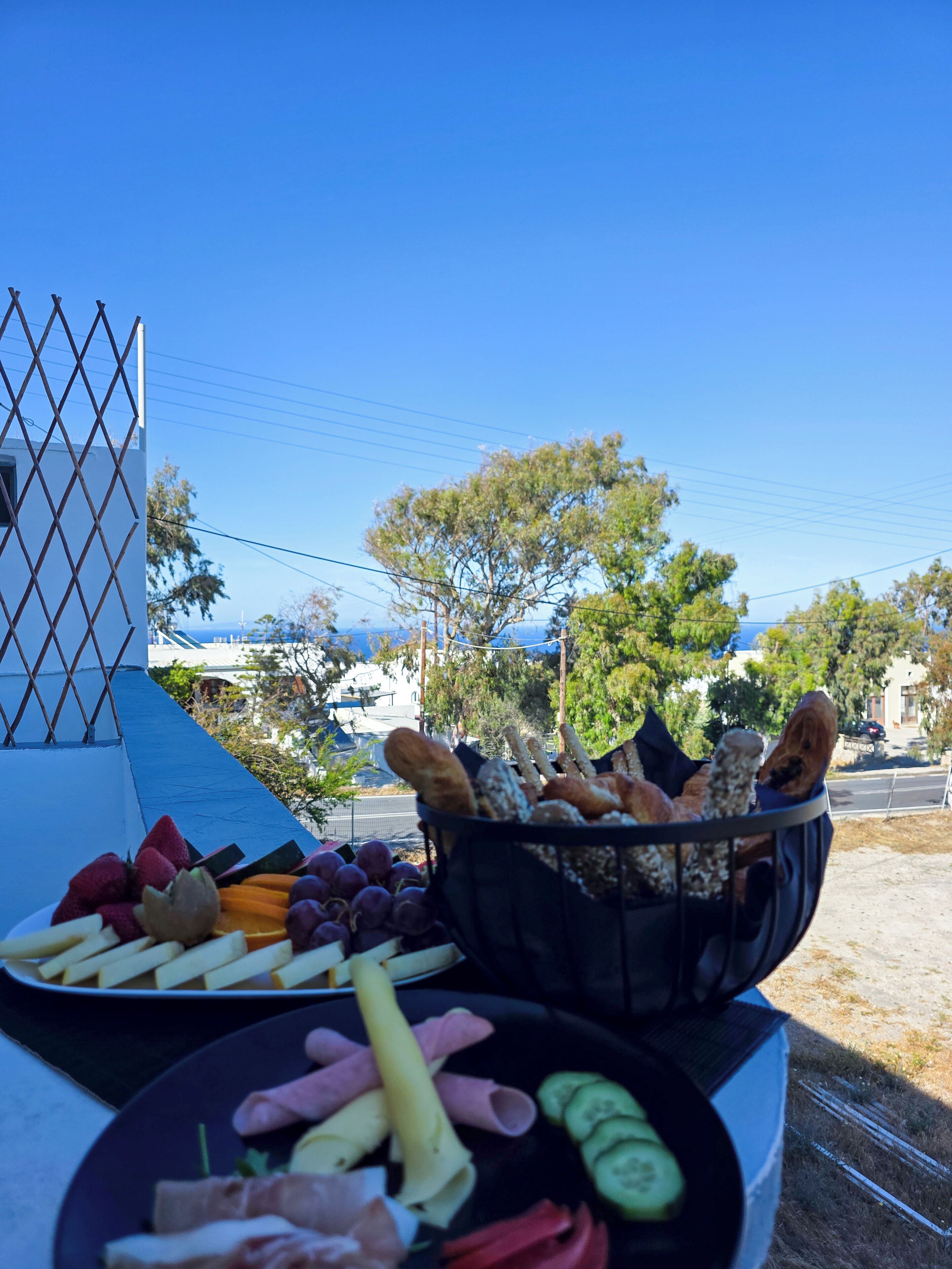 A table with a variety of snacks including grapes, cheese, and bread, set outdoors with a clear view of trees and buildings in the background.