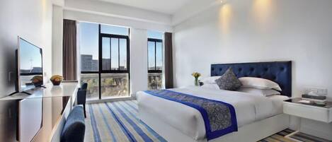 A hotel room with a large bed, a desk, a chair, a TV, and a balcony with a city view.