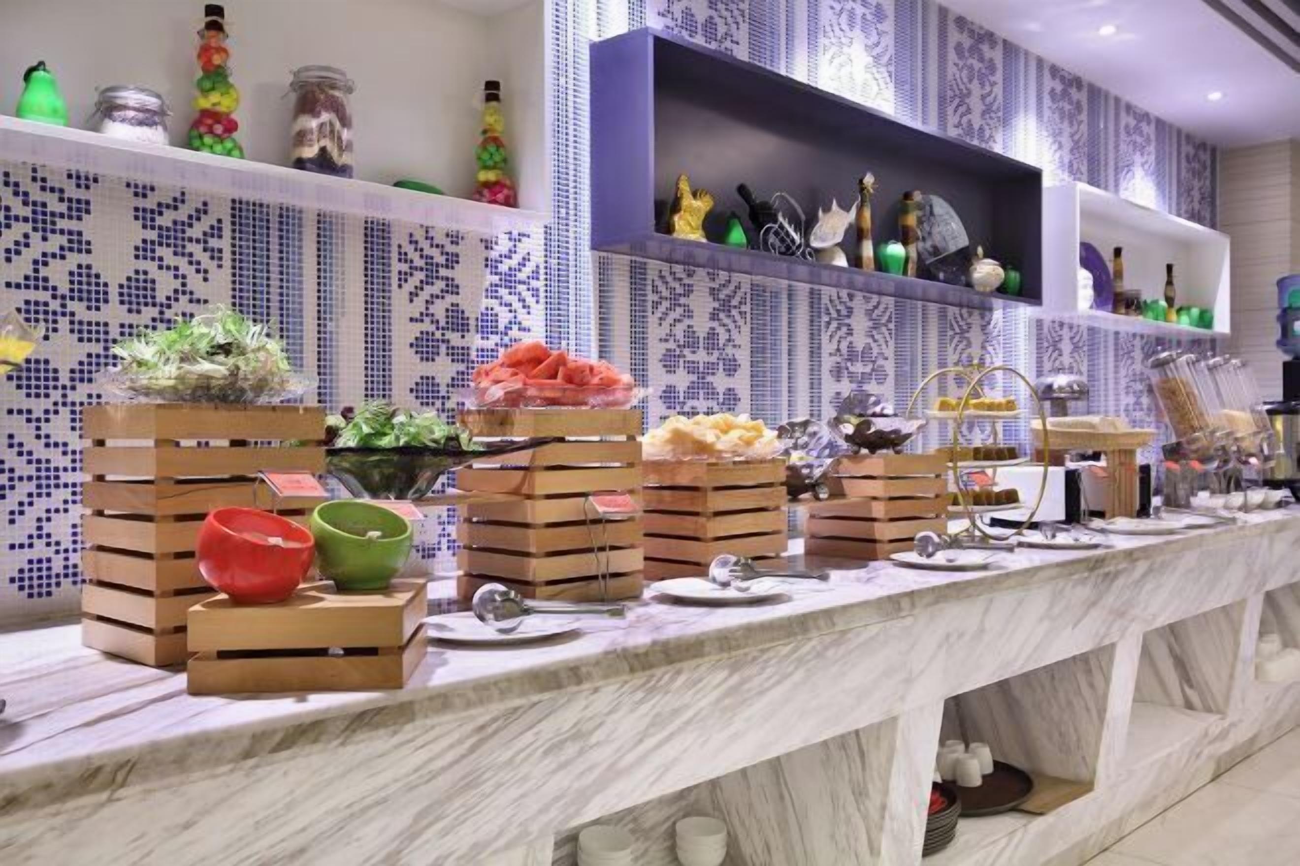 A buffet setup with various food items and utensils on a marble countertop, surrounded by patterned tile walls and shelves with decorative items.