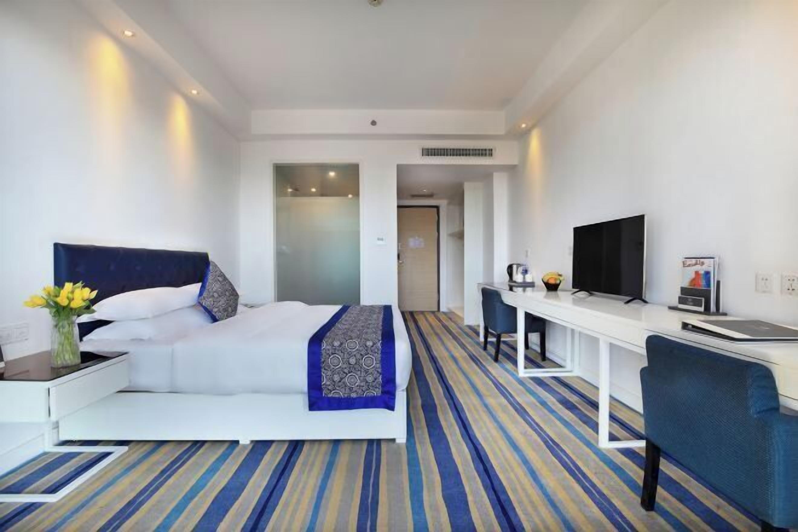A hotel room with a bed, a desk with a chair, a television, and a bathroom visible through a glass door.