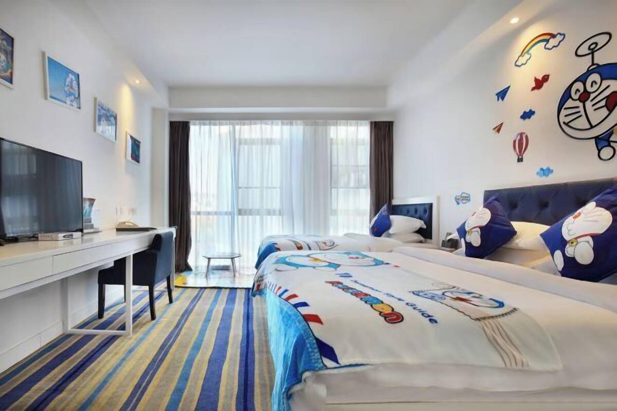 Habitación de hotel con una cama grande, un escritorio con televisor de pantalla plana, una ventana con cortinas y decoración en las paredes que incluye personajes de dibujos animados.