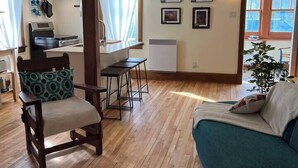 A cozy living room with a wooden floor, a green sofa, a wooden chair with a cushion, and a small table. There is a kitchen area with a stove, a refrigerator, and a window with curtains. - Retro-chic retreat in the heart of Wakefield (Wakefield)