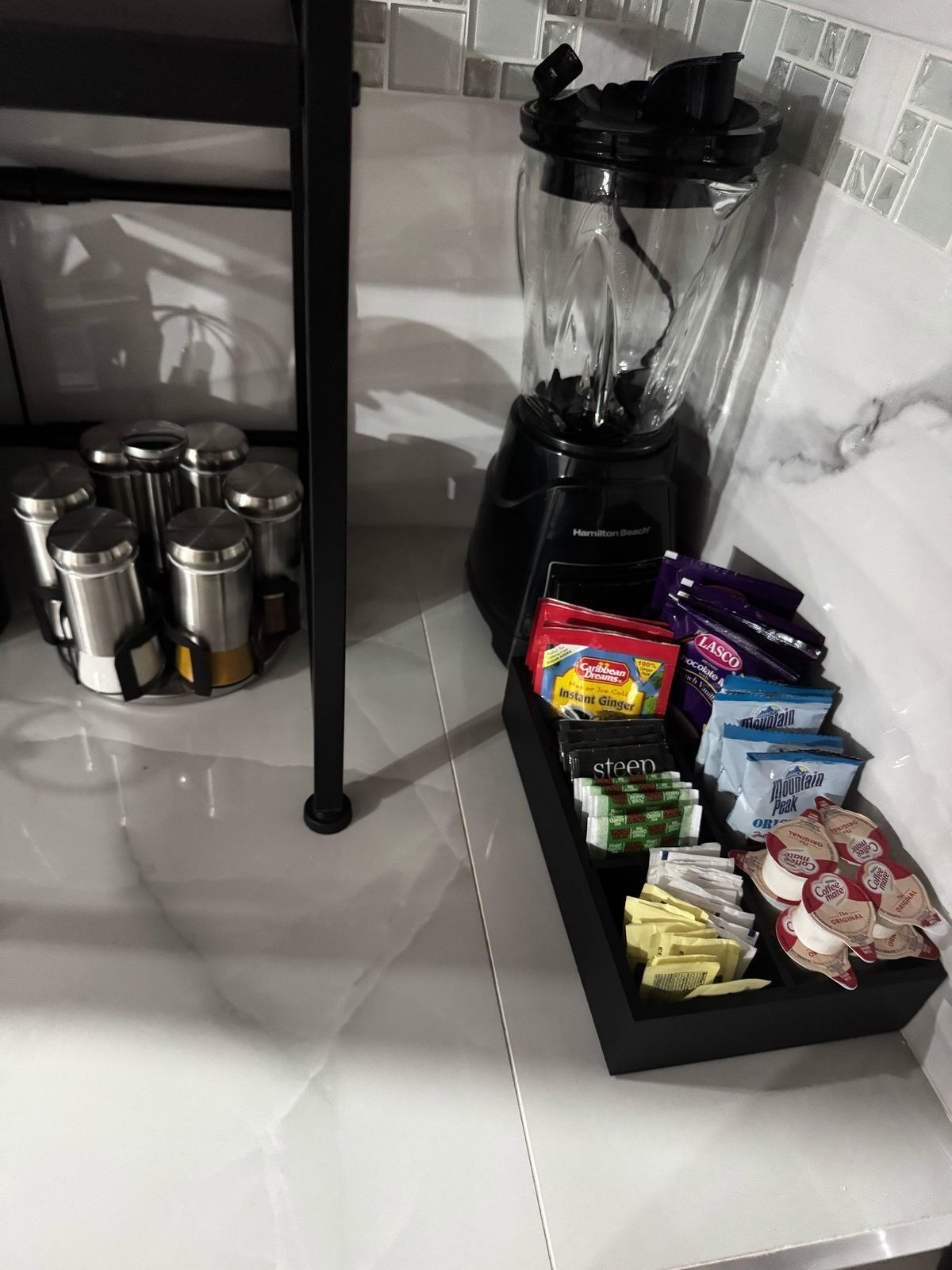 Un plan de travail de cuisine avec un mixeur, des récipients en acier inoxydable et divers aliments, notamment du café instantané et des en-cas.