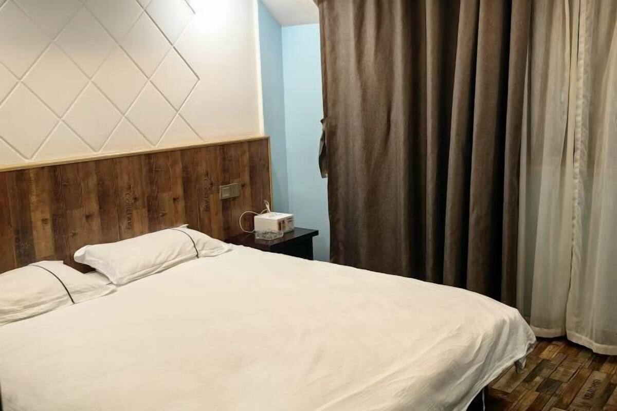 A hotel room with a wooden headboard, a bed with white bedding, a bedside table with a cup, and a curtain.