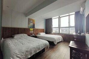 A hotel room with two beds, a wooden headboard, a large window with curtains, and a small table with a kettle.